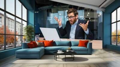 Angry and angry businessman talking on video call, talking to colleagues and shouting quarrel, mature boss working inside modern office building, senior man with laptop and beard. Wall mural