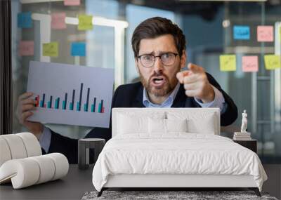 Angry businessman engages in video call, holds chart indicating displeasure during office presentation. Expression of frustration and urgency conveys stress in professional environment Wall mural