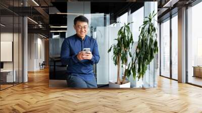 Asian businessman inside the office in a casual shirt and glasses is using the phone, the boss is holding a smartphone in his hands reading news and typing a message smiling. Wall mural