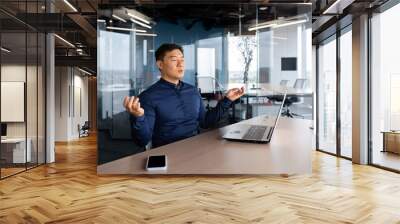 Asian businessman meditating in office, man working at desk using laptop, worker with closed eyes in lotus pose inside building. Wall mural