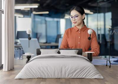 Asian businesswoman working on her laptop while taking notes in a modern office setting. She looks focused and professional. Wall mural