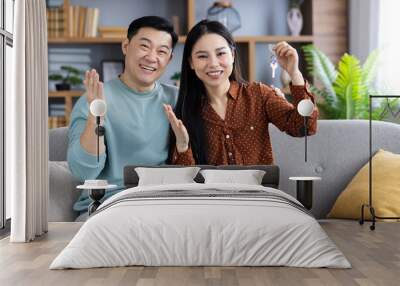 Asian couple joyfully presents new home keys while sitting on sofa in stylish living room, celebrating new beginnings and future together. Wall mural