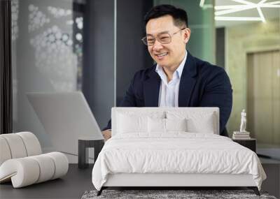 Asian mature man typing on a laptop in a stylish, modern office setting, demonstrating productivity and happiness at work. Wall mural