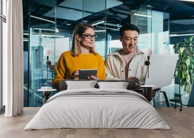 Business meeting of two employees, Asian and female colleagues, discussing the project, working in a modern office, looking at a laptop screen, discussing edits, and project success Wall mural