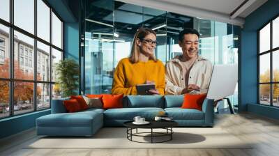 Business meeting of two employees, Asian and female colleagues, discussing the project, working in a modern office, looking at a laptop screen, discussing edits, and project success Wall mural