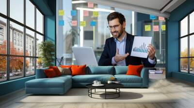 Businessman presenting data analysis in modern office setting. Smiling professional using laptop and charts to discuss growth. Concept of business strategy, success, and teamwork among office workers Wall mural