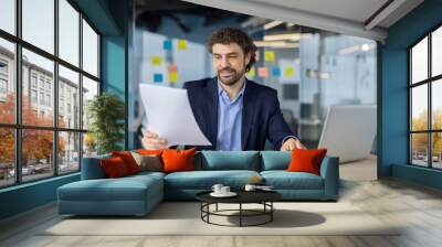 Businessman sitting at desk in modern office, reviewing documents and using laptop for work. Professional setting emphasizes productivity, focus, and engagement. Wall mural