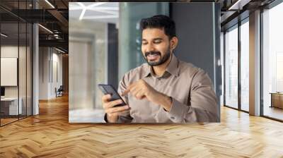 Careless employer having break from work while using mobile device on background of corporate building interior. Smiling man scrolling feed of social media for distracting from tensed project. Wall mural