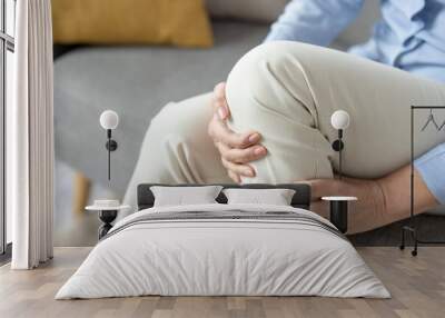 Close-up image of a mature woman sitting on a couch and holding her knee, depicting pain or discomfort in the joint. Wall mural