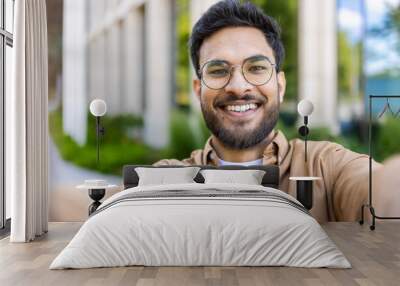 Close-up of smiling man taking selfie outdoors on sunny day. Man with beard and glasses in casual clothing enjoying outdoor moment. Concept of happiness, modern communication, and casual lifestyle. Wall mural