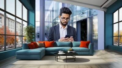 Close-up photo of a young Indian man in a business suit walking on the street near an office center and using a mobile phone. Wall mural