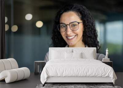 Close up photo portrait of beautiful Latin American woman with curly hair and glasses, businesswoman inside office building smiling and looking at camera Wall mural