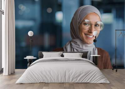 Close up photo portrait of beautiful young muslim woman, woman in hijab and glasses smiling and looking at camera, businesswoman working inside modern office building Wall mural