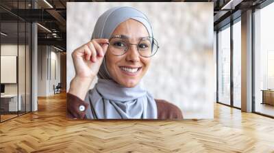 Close-up photo. Portrait of a young beautiful Muslim woman in a hijab smiling looking at the camera and adjusting her glasses with her hand. Wall mural