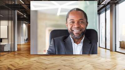 Close up portrait of successful successful businessman, man looking joyfully at camera sitting at desk inside office, satisfied african american boss. Wall mural