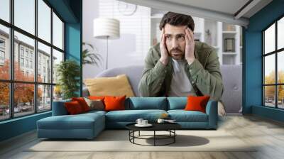 Concerned man seated on sofa holding head, depicting stress and anxiety in domestic setting. Middle-aged male wears casual attire, expressing worry and contemplation. Natural light illuminates the Wall mural