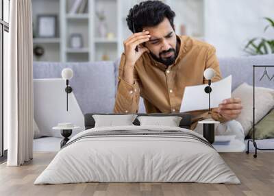 Concerned young Indian man sitting on a couch in home setting, examining papers with a laptop and calculator nearby. Wall mural