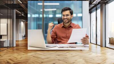 Confident businessman celebrates success at modern office desk with laptop and documents in hand, feeling accomplished during workday Wall mural