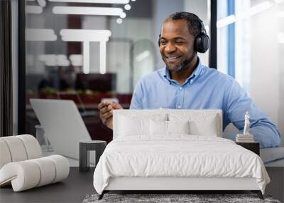 Confident businessman with headphones participates in virtual meeting using laptop and taking notes. Expressions of happiness and productivity reflect during online video conference in workspace. Wall mural