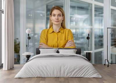 Confident businesswoman wearing a yellow shirt stands with arms crossed in a modern office setting, looking directly at the camera Wall mural