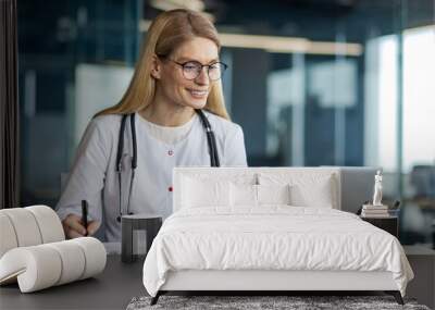 Confident female doctor wearing glasses and stethoscope using laptop in professional office setting. She is writing on paper, showcasing multitasking skills in medical industry. Wall mural