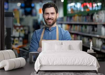 Confident male employee with apron stands with crossed arms in grocery store aisle, exuding professionalism and customer service expertise, surrounded by variety of products in retail setting. Wall mural