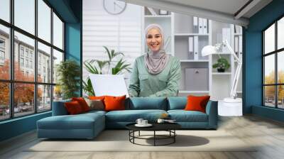 Confident professional Muslim woman in a hijab smiles at the camera while using her laptop in a modern office setting. Wall mural