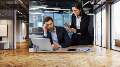 Conflicted office scene showing an angry female boss yelling at a frustrated male employee working on laptop. Tension highlights workplace stress and pressure during a business meeting. Wall mural