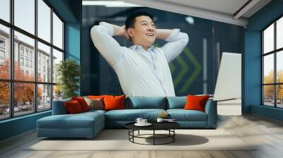 Dreamy successful Asian businessman working in office, smiling, hands behind head, thinking and building new dreams and plans. Wall mural