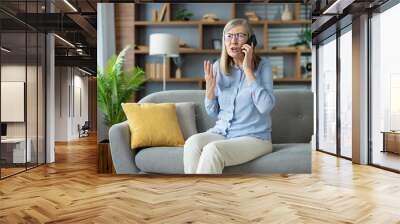 Elderly woman sitting on a couch, expressing frustration while talking on the phone with customer support in living room. Wall mural