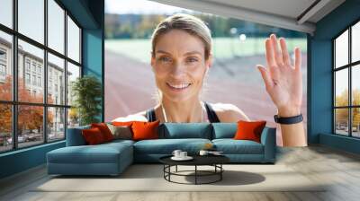 Female athlete after jogging in stadium talking with friends and recording online video blog, mature blonde with headphones looking at smartphone camera smiling, video call with headphones. Wall mural