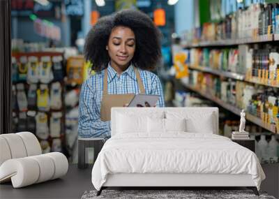 Female employee in a retail store aisle, using a tablet for inventory management. She wears a checkered shirt and an apron, focusing on digital tasks surrounded by various products. Wall mural