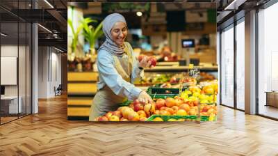 Female seller in hijab browsing and checking apples in supermarket, woman in apron smiling at work in store in fruit and vegetable department. Wall mural