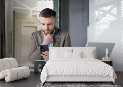 Focused businessman using smartphone at modern office desk with laptop, conveying urgency, tech, and professional lifestyle. Wall mural