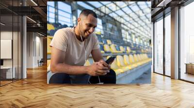 Happy and satisfied man after jogging and fitness class sitting and using smartphone, African American man in headphones listening to online podcasts, audio books and music, typing on phone Wall mural