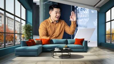 Happy and successful asian boss talking with colleagues on video call, using laptop, waving hands greeting to camera, businessman at work Wall mural