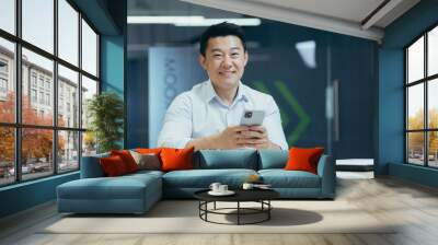 Happy Asian man using phone in office, businessman smiling and looking at camera typing message and browsing internet page Wall mural