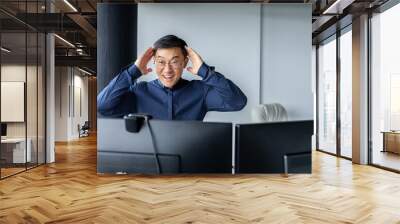 Happy business broker looking at two paired computer monitors and happy, businessman working inside modern office building, asian man celebrating successful deal and signed contract. Wall mural