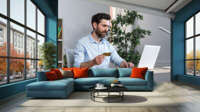 Happy businessman makes online purchases working in the office, husband holds credit bank card and uses laptop Wall mural