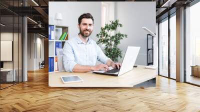 Happy businessman working in bright modern office behind laptop man with beard smiling and looking at cameras Wall mural