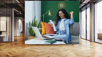 Happy hispanic woman at home doing paperwork and calculating household budget, sitting on couch in living room and using laptop at work, holding hand up victory and triumph gesture. Wall mural