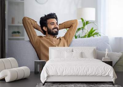 Happy Indian young man sitting relaxed on sofa at home with hands behind head and resting, smiling contentedly. Wall mural