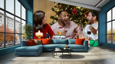 Happy young interracial people celebrating new year at home at festive table near Christmas tree. They hold glasses in their hands, laugh, communicate, rejoice. Wall mural