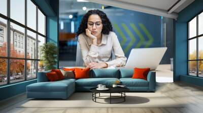 Hispanic businesswoman looking exhausted working on laptop in office. Surrounded by crumpled papers and documents, expressing stress and fatigue. Concept of overwork, deadline pressure Wall mural