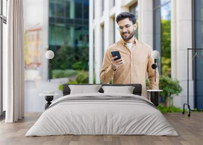 Hispanic man in casual attire standing outside modern office building using smartphone. Daylight, urban setting, technology and business concepts. Wall mural