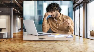 Indian businessman in casual shirt appearing stressed, sitting at his office desk with laptop, holding glasses in his hand. Wall mural