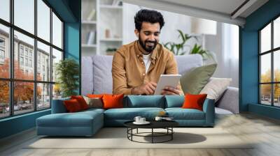 Indian man smiles while browsing on a tablet, comfortably seated on a sofa in a well-lit modern apartment living room. Wall mural