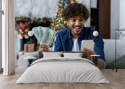 Man celebrating victory and success sitting on sofa at home in living room near Christmas tree. Hispanic man holds phone and money cash dollars in hands. A winner for the New Year holidays and Wall mural