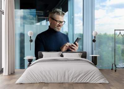 Mature adult man inside office standing near window, businessman typing message on phone and browsing internet pages, gray-haired boss smiling reading news online. Wall mural