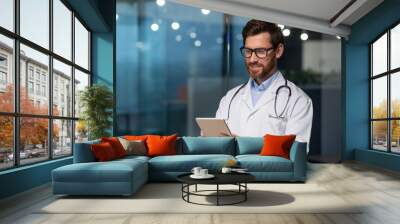 Mature and successful doctor in medical white coat and stethoscope working in boring clinic office, man in glasses smiling and using tablet computer, for online patient consultation. Wall mural